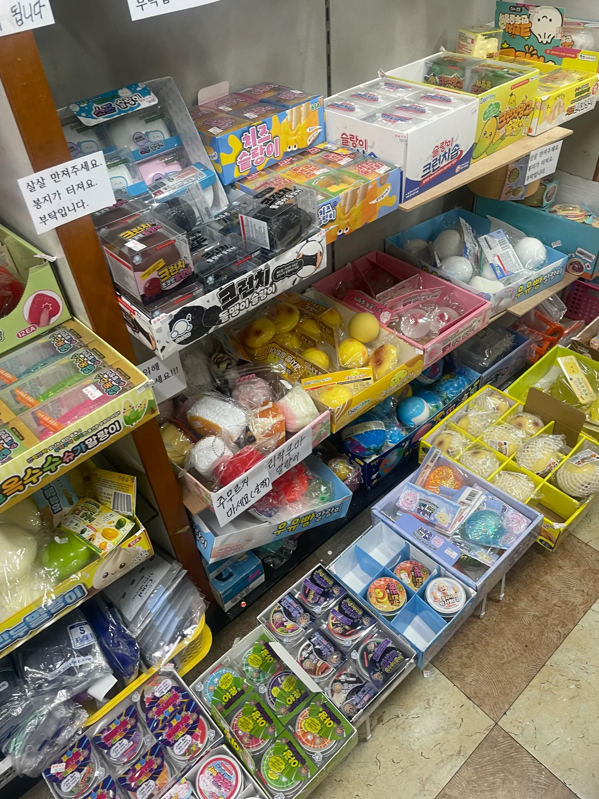 A vibrant display of various Korean Squishy toys, known as Mallang-ee, packed on shelves in a local Seoul shop for ASMR and stress relief. The collection features popular food-shaped items like milk bread, corn, and cheese squishies, alongside viral wax crushing sets, complete with handwritten Korean signs advising customers to handle them gently.