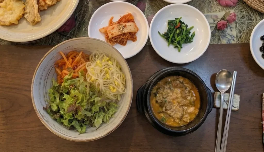 An aesthetic overhead view of a plant-based dining experience in a Seoul vegan restaurant, showcasing a bowl of bibimbap, soybean paste stew, and a variety of seasonal banchan.