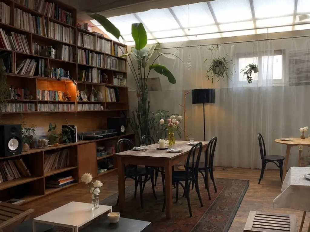 A cozy 2:3 vertical interior of a trendy gluten free cafe seoul with a large wooden bookshelf, indoor plants, and a 4-person wooden dining table with black chairs on a vintage rug. This aesthetic spot resembles a popular vegan cafes in seoul and a seoul vegan bakery, known for serving organic vegan desserts in seoul, sometimes referred to by K-pop fans as a jennie blackpink bakery style hangout.