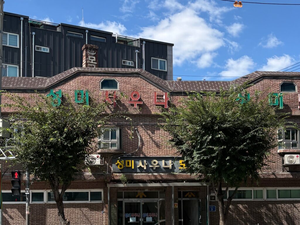 Korean spa exterior in Seoul with a retro brick facade, colorful Korean signage, and a classic neighborhood bathhouse vibe