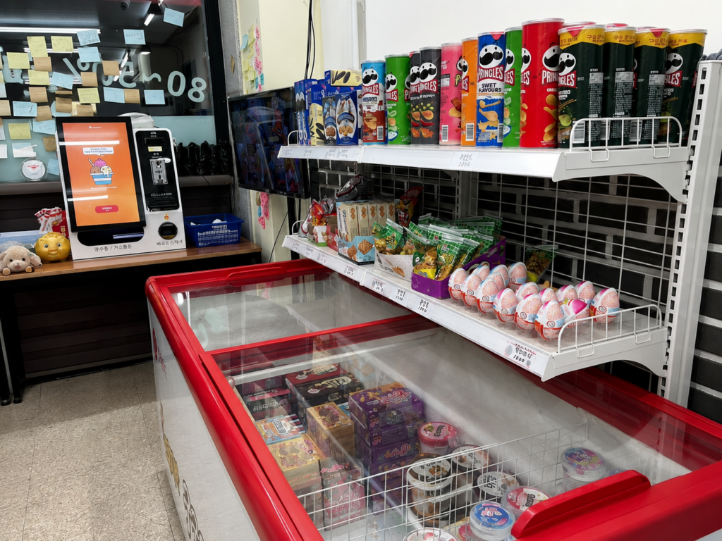 Korea unmanned convenience store with a snack shelf, colorful Pringles cans, Korean snacks, ice cream freezer, and self-service payment kiosk